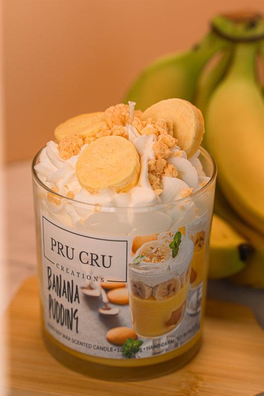 A jar candle with a label indicating it is a banana pudding scent, topped with a realistic depiction of bananas and cream.