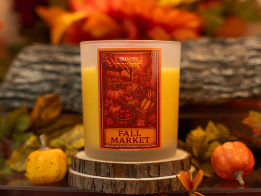 Candle with a 'Fall Market' label on a wooden surface with autumn decorations.