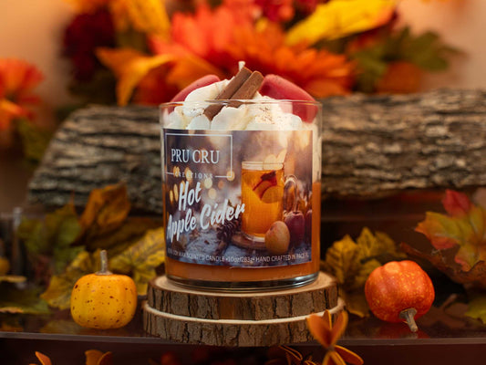 Pru Cru Hot Apple Cider candle on a wooden stand with fall-themed decorations.