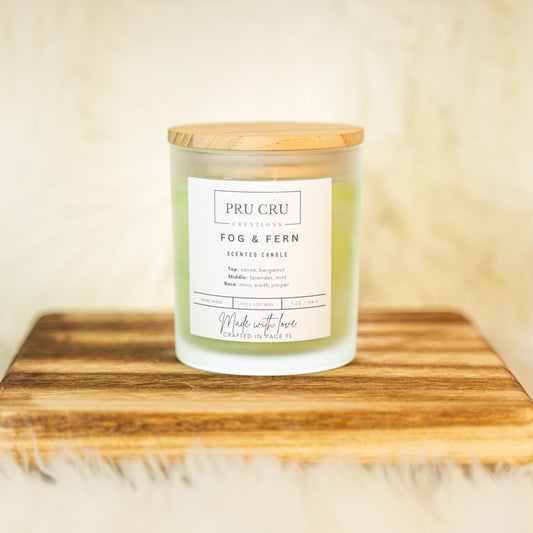 Candle labeled 'Pru Cru Fog & Fern' on a wooden surface with a soft background