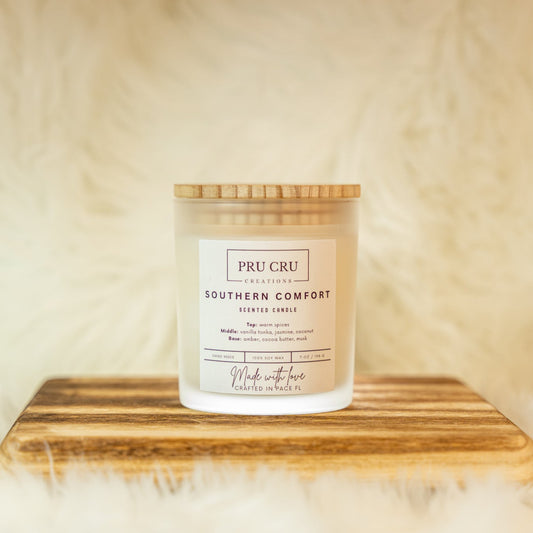 Candle labeled 'Pru Cru Southern Comfort' on a wooden stand with a soft beige background